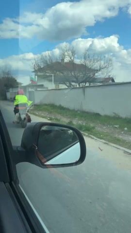 The video was captured from inside a car and shows a man riding an electric scooter. The man is wearing a red helmet, sunglasses, a bright yellow safety vest over a black vest, and black pants with the word "Regatta" written on one leg. He has a white bag with "JEFTINOC 45 kg UCI ELAGEL" printed on it resting on the scooter's floorboard. Dangling from the back of the scooter is a dead chicken. The scooter is black and has wide tires. The video follows the man for a few seconds as the car drives past. The scene takes place on a paved road in a residential area with houses, walls, and trees visible in the background under a partly cloudy sky.