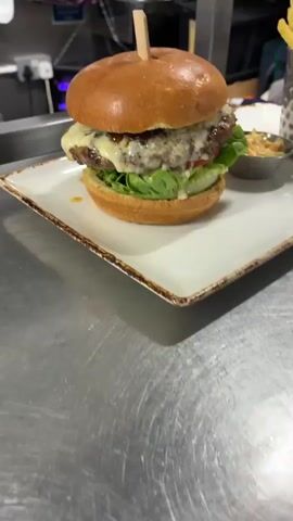 A close-up shot shows a gourmet burger on a white plate. The burger is large, with a glossy bun, a thick patty, melted cheese, lettuce, and tomato. A wooden skewer holds the top bun in place. To the right of the burger, a small dish of coleslaw is visible. The plate sits on a stainless steel surface, likely a kitchen counter. The background is blurred, but appears to be a commercial kitchen environment with some equipment and shelves. The lighting is bright and focused on the burger.