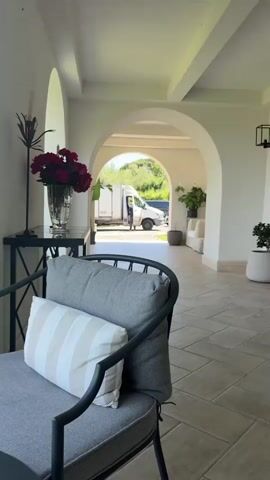 The video begins with a view of the interior of a building. There is a gray chair with a striped pillow in the foreground, a black table with a vase of flowers on top, and a series of arches leading to an outdoor space. Through the arches, there is a white van with a person standing near it. The camera then pans to the left, revealing a lush green lawn, trees, and a body of water in the background. The sky is blue with some clouds.