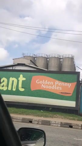 The video shows a roadside view of an industrial area. A large green billboard is visible, featuring the text "Golden Penny Noodles" on an orange banner. To the left of the banner, large yellow lettering is partially obscured. Above the billboard, industrial silos and structures are present. The scene is captured from inside a vehicle, as indicated by the reflection of the interior in a side mirror. The road surface and a sidewalk with grass are visible in the foreground. The sky is cloudy. There are no clearly visible people or specific actions being performed.