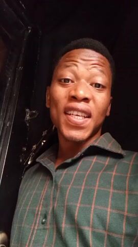 The video shows a man facing the camera and speaking. He has short, dark hair and is wearing a green and red plaid shirt. He is standing in what appears to be a doorway, with a black wall behind him. He says "Bismillah tawakaltu 'ala Allah".