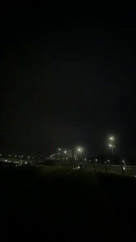 The video shows a dark nighttime scene with a street or highway illuminated by streetlights. The lights cast a yellow glow on the road and surrounding area. In the distance, there are more lights, suggesting a town or city. The overall atmosphere is quiet and still, with no visible movement or activity. The camera seems to be positioned on an elevated vantage point, overlooking the road.