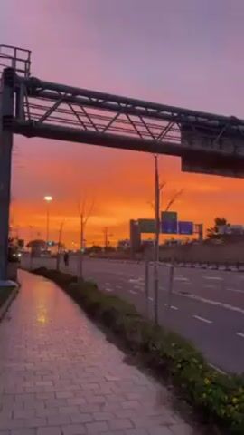 The video presents a vivid sunset scene adjacent to a roadway. The sky displays a dramatic gradient, shifting from deep purple overhead to a fiery orange near the horizon. An overhead metal structure, likely a sign support, frames the view. The roadway has multiple lanes with intermittent traffic, including cars and a cyclist. A brick sidewalk runs along the side of the road, bordered by greenery. Streetlights illuminate the area. Signage is visible on the road indicating directions or destinations. The overall environment suggests a suburban or urban setting during dusk or twilight.