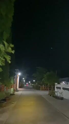 The video shows a dark, nighttime street scene. A paved road stretches forward, illuminated by streetlights at intervals. On the left side of the road, there is a building with a bright light above its entrance, and some potted plants. On the right side of the road, there are trees and some wooden structures. A single red motorcycle is visible in the distance, traveling away from the camera. The sky is dark and appears clear, with a few faint objects that could be stars or distant lights. The overall atmosphere is quiet and somewhat serene.
