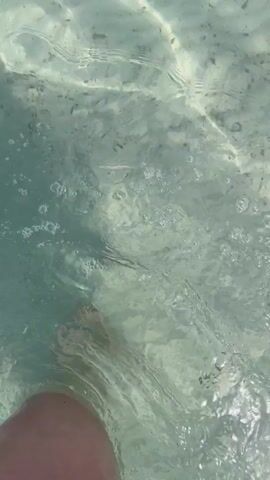 A first-person perspective video shows a person's legs and feet submerged in clear, shallow water. The camera is underwater, looking up towards the surface. Sunlight filters through the water, creating shimmering patterns on the sandy bottom. Small fish can be seen swimming in the background. The person is moving their legs, causing ripples and splashes on the water's surface. The video appears to have been taken during the daytime.
