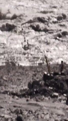 A drone is hovering over a turbulent, muddy river. A person is standing on a rocky, debris-strewn bank, operating a small yellow crane-like device. The drone is equipped with a cage-like attachment.
