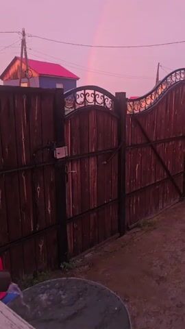 The video shows a wooden gate with ornate metalwork in front of a rural setting. A faint rainbow is visible in the pale pink sky, suggesting it might be dawn or dusk. Two houses with red roofs are visible behind the gate, along with utility poles. The ground in front of the gate is dirt, and a round, dark object, possibly a table or a barrel, is in the foreground. The overall mood is tranquil and serene, enhanced by the soft colors of the sky and the natural elements. The location is identified as Khatassy, Russia.