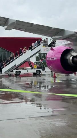 The video shows passengers boarding a Wizz Air plane on a wet tarmac. A mobile staircase is positioned at the plane's entrance, and people are walking up the steps to board. The plane has a white body with pink engine covers. A section of the airport building, with a red facade, is visible behind the plane. The ground is shiny and wet, reflecting the overcast sky. A green safety line lies on the ground. The scene appears to be at an airport during or after a rainfall.