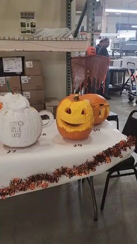 A table covered in a white tablecloth displays several Halloween pumpkins. The table is decorated with orange and black garland. The pumpkins are carved with various designs, including faces, a cat, and a pumpkin spice latte. Numbers are placed in front of each pumpkin. Shelves full of boxes are in the background. Two people are visible in the background.