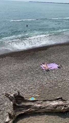 A person in a bikini lies on a purple towel on a rocky beach, facing away from the camera. Their arms are spread wide. In the background, the ocean is calm with small waves, and a person can be seen swimming. A large piece of driftwood is in the foreground, partially submerged in the water. The sky is overcast.