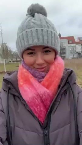 A woman in a grey puffy jacket, a grey beanie with a pompom, and a pink and orange scarf smiles at the camera. She is outside on an overcast day, in what appears to be a grassy field with some trees in the background. Buildings are visible in the distance. The camera seems to be held at arm's length for a selfie perspective, and there is a slight shaking motion to the video.
