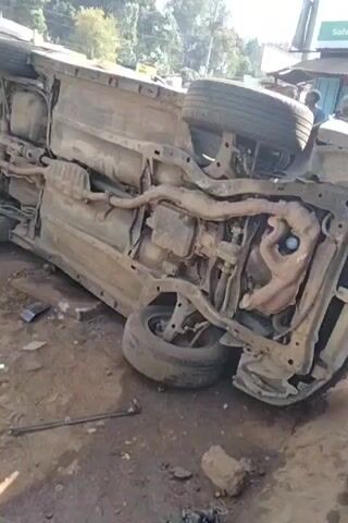 The video shows the aftermath of an accident. A car is overturned, revealing its undercarriage, including the tires, exhaust system, and other mechanical components. Debris is scattered around the vehicle. Next to it, another vehicle, possibly a small truck, is also overturned. A tow truck is visible in the background. A few people are standing nearby, seemingly observing the scene. The setting appears to be an outdoor area next to a building or structure with a sign that says "Safaricom" on it. The ground is dirt, with scattered pieces of brick and other debris. The overall impression is one of a recent and serious accident.