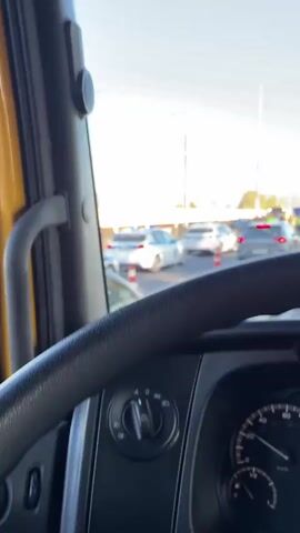 This is a first-person view from the cab of a vehicle, likely a truck, looking out the windshield. The camera pans slightly to the right, showing a traffic scene. In the foreground, the steering wheel and part of the dashboard are visible. The road ahead is congested with vehicles. Two police cars are parked on the left side of the road, their lights flashing. Further ahead, there is a significant car accident. A severely damaged dark-colored car is in the middle of the road, with its front end crumpled. Several people are standing around the accident scene, some appearing to be police officers and others possibly emergency personnel or witnesses. One individual in a high-visibility vest is directing traffic. Traffic cones are placed around the accident area. In the background, there are power lines and hills under a clear blue sky. The driver's side mirror is visible, reflecting the scene behind, including the police cars and traffic.