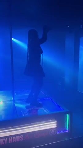 A person is silhouetted on a raised, illuminated platform in a dimly lit environment. The platform has a mirrored surface and is outlined with colorful LED lights. The person, with long hair, is in a dynamic pose, seemingly dancing or performing. The background features a vertical pole and beams of colored light, predominantly green and blue, creating a club-like atmosphere. Text on the lower part of the platform reads "FUNKY HAUS It's a Lifestyle". The video cuts between different lighting conditions, with the colors shifting from green to blue and purple. In one brief shot, another person wearing a cap is visible in the background to the left.