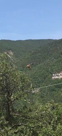 A helicopter hovers in the air, suspended above a lush, green, mountainous landscape. The helicopter is orange and has a long cable attached to it, which trails down to a white bucket. The bucket appears to be suspended from the cable. The sky is blue with a few wispy clouds. The helicopter is in the process of lifting or lowering the bucket.