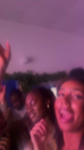 The video captures a close-up shot of a woman with dark hair, wearing a black top, who is singing enthusiastically and excitedly at a party. The lighting is predominantly pink, possibly from neon lights or a similar source, creating a vibrant, party atmosphere.  In the background, other people can be seen, suggesting a crowded environment. One individual is visible behind and to the left of the main subject, adding to the sense of being in a busy social gathering. The camera is somewhat shaky, likely hand held, which adds to the feeling of movement and energy typical of a party setting.