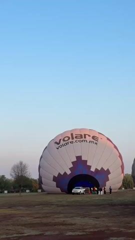 The video shows hot air balloons being prepared in a field. In the foreground, there is a partially inflated balloon with the word "Volare" and "volare.com.mx" printed on it. A white truck is parked near the opening of the balloon, and several people are standing around. In the background, two other hot air balloons are visible in the sky, one higher than the other. The sky is a clear blue, and there are some trees and a building visible in the distance.