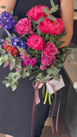 A person dressed in a black dress holds a bouquet of flowers. The bouquet contains bright pink peonies, smaller pink roses, purple delphiniums, and other colorful flowers and greenery. The bouquet is tied with a pink ribbon, and a small white tag with the letter "B" printed on it is attached. The background appears to be an indoor setting with a gray surface and a hint of a wall. The lighting is bright and natural, illuminating the vibrant colors of the flowers.