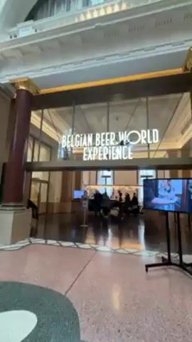 The video shows the interior of a building with high, ornately decorated ceilings and large windows. The camera pans upward to showcase the architecture, which includes columns, arches, and a skylight. The entrance to the "Belgian Beer World Experience" is visible, with people inside. A green sign is also present.