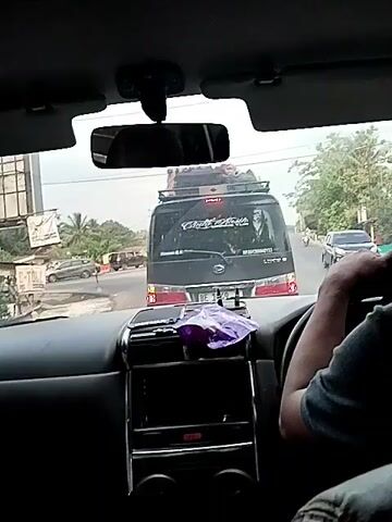 The video begins inside a vehicle, looking out through the windshield. In front of the vehicle is a black van with luggage strapped to the roof. The van has writing on the back window and a picture of a person. A license plate is visible on the van. Further ahead, there are other vehicles and a motorcycle. The environment appears to be a road in a tropical or subtropical region, with trees and vegetation visible on the sides. A hand, presumably the driver's, is visible on the steering wheel. The scene suggests a traffic situation, possibly a traffic jam or a slow-moving road. The camera is stationary, capturing the view from the passenger seat.