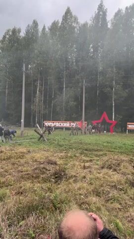 A group of people in military-style clothing, some carrying red flags and what appear to be rifles, are marching in formation through a grassy field. Behind them is a line of trees and a banner with Russian text. To the right of the formation is a large red star. In the foreground, a person's head and shoulders are visible, suggesting they are filming the event. The overall atmosphere appears to be a reenactment or ceremony.