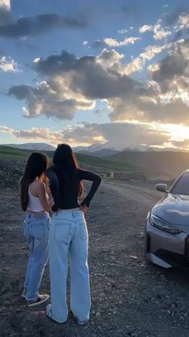 Two women are standing on a dirt road next to a gray BMW. The car's license plate reads "66 MH 066" and appears to be Armenian. The women are facing away from the camera, looking out at a valley with rolling green hills and a snow-capped mountain range in the distance. The sun is setting, casting a warm glow on the scene and creating dramatic clouds in the sky.