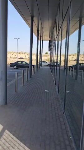 A low-angle, stationary shot shows a paved walkway running alongside a modern building with a reflective glass facade. The walkway is bordered by sleek, cylindrical metal bollards and large, grey pillars supporting an overhang. The glass exterior reflects the sky, clouds, and the surrounding environment, creating a sense of depth.

On the left side of the frame, a parking lot is visible with several dark-colored sedans. Beyond the parking lot, a low, arid landscape with sandy hills and sparse vegetation suggests a dry, possibly desert-like environment. A few streetlights are also present. The scene appears to be outdoors, under a clear, bright sky, indicating daytime with possibly warm weather. The lighting suggests it's either morning or late afternoon.

No people are visible in the frame. The focus is on the architecture and the reflections in the glass. The overall impression is one of a contemporary, functional space, perhaps an office building or a commercial establishment, situated in a semi-arid region. The repetition of the pillars and the reflections in the glass creates a sense of order and symmetry.

There is some faint text visible on a blue sign reflected in the glass, but it is too blurred to be read. Another item of text on what appears to be a discarded flyer or pamphlet is visible on the walkway.