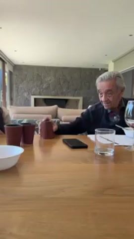An elderly man with gray hair is sitting at a wooden table, rolling dice using a leather dice cup. He is wearing a dark-colored sweater and has a pair of glasses hanging around his neck. On the table, there is a smartphone, a glass of water, a wine glass, a white bowl, and another leather dice cup. A younger person is partially visible to the left of the frame. The room features a stone fireplace in the background, and the ambiance is bright and well-lit.