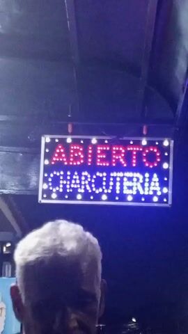 A sign reading "ABIERTO CHARCUTERIA" in red and blue LED lights hangs above a street at night. The sign is rectangular with a white frame. To the left, a menu is displayed with the word "CHARCUTERIA" at the top, followed by a list of items in Spanish. The street is lined with streetlights, and several people walk along the sidewalk in the background.