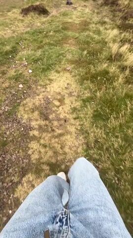 A first-person perspective video shows a person sitting on a grassy slope. The camera is pointed downwards at the person's legs, which are clad in blue jeans and white sneakers. The person is facing a pathway that leads uphill. In the distance, a person is standing on the pathway. The ground is covered in grass and patches of brown dirt.