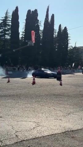 A dark blue car is drifting, creating a large cloud of smoke, on a paved asphalt surface. Several orange traffic cones are strategically placed around the car on the road. In the background, a line of tall, dark green trees stands against a clear blue sky. Beyond the trees, there are buildings and what appears to be a crowd of spectators. A red flag or banner is visible hanging from a pole in the mid-ground. The scene suggests an outdoor event, possibly a car show or competition.