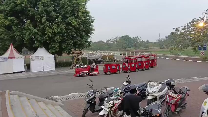 The video begins with a wide shot of an outdoor scene, likely a park or recreational area, as indicated by the presence of trees, a paved pathway, and what appears to be a body of water in the background. In the foreground, several motorcycles are parked. A set of stairs leads up to a paved area where a miniature red train is positioned. The train consists of a locomotive and several passenger cars, all painted red. A person is visible in the locomotive. To the right of the train, there are several tents, one of which is red and white striped, and others are white. Behind the train and tents, a large golden statue of a robot, resembling a Transformer, stands prominently. A bridge spans across the body of water in the distance. The sky is overcast. The camera pans slightly to the right, revealing more of the parked motorcycles and the general layout of the area. The train begins to move slowly forward, with the locomotive and its attached cars traveling along the paved path. The scene appears calm and possibly set up for visitors, with the tents suggesting vendor stalls or information booths. The overall impression is a leisurely outdoor setting with attractions for visitors.