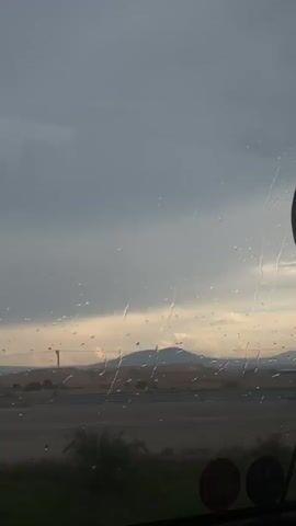 The video, filmed from inside a vehicle, shows raindrops on a window. Outside, a yellow and black chevron sign indicating a curve to the right is visible. Beyond the sign, a road stretches towards a distant mountain range under a cloudy sky. The time stamp indicates the video is approximately 3 seconds long.