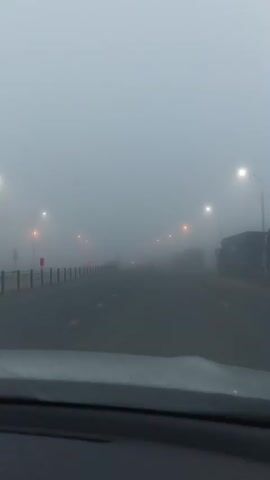The video shows a view from inside a vehicle driving on a road in heavy fog. The road ahead is visible, with streetlights lining both sides. There are other vehicles on the road, including cars and trucks, which are difficult to see due to the fog. The fog obscures the surroundings, creating a limited visibility environment. The overall scene is dominated by the thick fog, which lends a muted and ethereal quality to the footage.