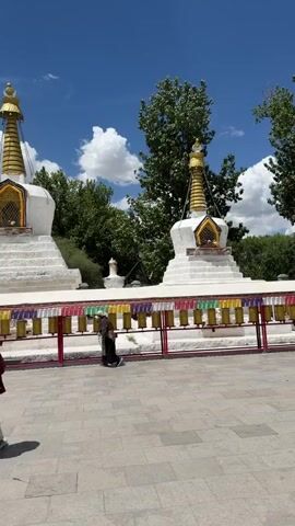 The video captures an outdoor scene on a bright, sunny day with a clear blue sky and some white clouds. In the foreground, several people are walking across a paved area from left to right. One person in the middle of the frame, dressed in dark clothing, appears to be engaging with a row of colorful prayer wheels, possibly spinning them.

Dominating the background are two large white stupas with tiered bases and ornate golden spires, characteristic of Buddhist architecture. They are positioned behind the prayer wheels. Beyond the stupas, lush green trees are visible, and in the distance, a hazy mountain range can be seen. The overall setting suggests a temple or monastery grounds in Chengguan District, China. The atmosphere appears peaceful and calm.