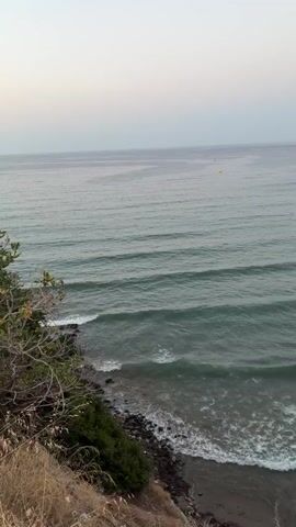 The video shows a wide, calm ocean with gentle waves rolling towards a rocky and vegetated shoreline. Several people are visible swimming or floating in the water at varying distances from the shore. The sky is clear with a soft gradient of color. A yellow buoy is visible in the distance. The camera pans slightly to the right, revealing a more expansive view of the coastline with a large hill or mountain in the background.
