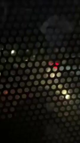 The video shows a view through a screen of small holes, obscuring the outside.  Red and white lights are visible, likely from vehicles at night. The view is shaky, suggesting movement or instability of the camera. There are indistinct shapes outside, possibly buildings or other objects, but their details are unclear due to the screen and low light.