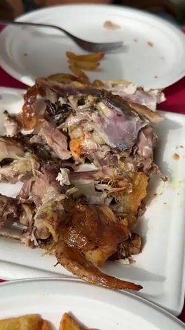 A close-up, top-down view of the remains of a roasted chicken on a white rectangular plate. The meat has been picked clean, leaving behind bones, skin, and some scraps of meat. A fork is visible in the background, resting on another white plate that contains a few french fries. Two pairs of hands are present, one of which is using the fork to pick at the chicken carcass. The environment appears to be a table setting, with a red tablecloth visible beneath the plates.