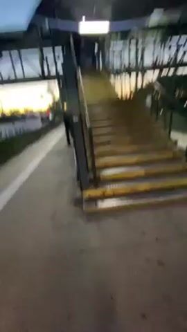A person walks up a set of metal stairs, as seen from a first-person perspective. The stairs are gray metal with yellow edges. The person wears black pants and off-white sneakers. The concrete floor is visible at the bottom of the stairs. The lighting appears dim.