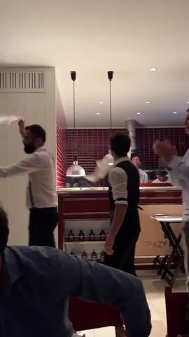 A group of people are shown dancing and celebrating indoors. Several individuals are wearing formal attire, including white shirts, bow ties, and suspenders, suggesting a restaurant or catering staff. They are waving white napkins in the air and appear to be engaged in a lively, celebratory dance. The environment appears to be a restaurant with ambient lighting and a visible kitchen area in the background. The overall atmosphere is festive and energetic.