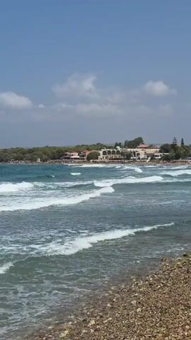 This is a vertical video of a beach on a sunny day. The water is a deep blue with white waves crashing onto a pebbly shore in the foreground. In the mid-ground, there is a distant shoreline with trees and buildings. The sky is a clear blue with a few scattered white clouds. The camera is stationary.