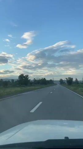 This is a first-person perspective video taken from a vehicle driving on a straight asphalt road. The scene is rural, with green fields and scattered trees lining both sides of the road. In the distance, there are rolling hills, and several power lines and pylons are visible, suggesting the presence of infrastructure. The sky is a vibrant blue with scattered, fluffy white clouds, indicative of a fair-weather day. The sun appears to be low in the sky, casting a warm light and suggesting it is either early morning or late afternoon. There is no text visible in the video. The overall mood is peaceful and suggests a journey or travel through the countryside.
