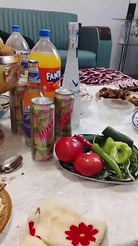 The video shows a table laden with food and beverages. A large, decorative gold bowl overflows with fruit, including apples, bananas, and grapes. There are several bottles and cans of drinks, including Fanta, Tosee, and Moxito. A bottle of Grey Goose vodka is also visible. Plates of food are scattered around the table, including one with meat and another with fresh vegetables like tomatoes, cucumbers, bell peppers, and green peppers. Spoons and small bowls are set out, and a small white cloth with a red flower pattern lies near the front of the table. The background features a turquoise couch and other furniture in what appears to be a living room. Loud music is playing in the background.
