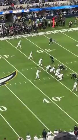 The video shows a football game from an elevated position, likely within a stadium. Two teams, one in white uniforms and the other in dark uniforms, are facing each other on a green field marked with yard lines. The white team is in possession of the ball, and the quarterback is attempting a pass. Several players from both teams are actively engaged, either blocking or trying to intercept the pass. The background reveals a stadium packed with spectators. The lighting appears bright, indicating it is likely a daytime game. The camera angle is slightly angled, providing a view of a significant portion of the playing field.