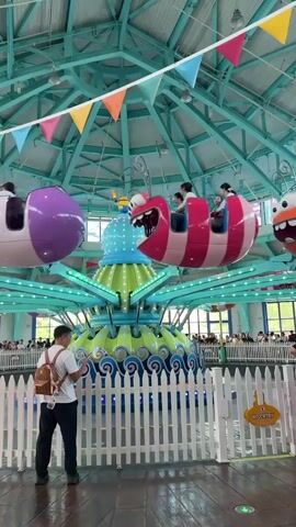 A man stands in front of a white picket fence, facing away from the camera, holding a smartphone. He is wearing a white t-shirt, dark pants, and a brown backpack. Behind him, a circular amusement park ride with cartoonish creatures is in motion. The ride features several hanging pods, one pink and white, another orange and white, and a purple one. The central part of the ride is green and blue, decorated with wave-like patterns. Colorful triangular flags are strung across the ceiling above the ride. Several people are visible on the ride and in the background. The environment appears to be indoors, with a high ceiling and windows visible in the distance.
