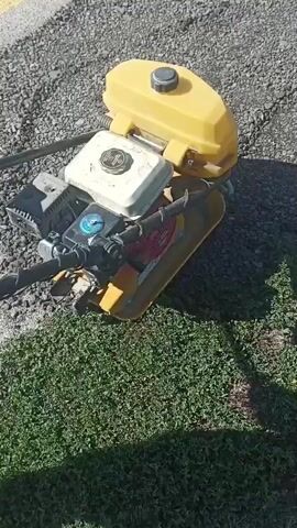 A person wearing black shorts, a green shirt, and black shoes is operating a yellow and white plate compactor on a surface of gravel and grass. The plate compactor has a yellow fuel tank and a white engine. The person is holding the handles of the plate compactor, and the machine is vibrating. The ground is covered with small gray pebbles. There is also green grass in the foreground. The sun is shining, casting shadows on the ground.