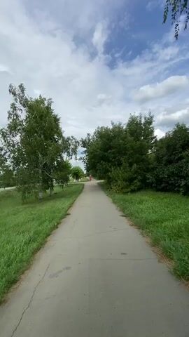 This is a selfie-style video taken outdoors on a cloudy day. The primary subject is a young woman wearing a white long-sleeved shirt, a white baseball cap with an embroidered logo, and black pants. She is walking along a paved walkway. To her left is a grassy area, and behind her, further away, are some buildings with green roofs, reminiscent of school or institutional architecture, and some trees. The overall setting appears to be a somewhat urban or semi-urban environment, possibly a park or a street in Akyar, Russia, given the provided location context. The woman is smiling slightly at the camera, suggesting a relaxed or pleasant mood. The sky is partly cloudy, indicating it is daytime. There is no significant action or event taking place other than the woman walking and filming herself. The logo on her cap appears to be an embroidered initial, possibly "M".