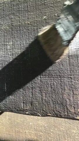 A close-up shot shows a paintbrush with black bristles being used to apply a dark substance to a textured surface that appears to be wood. A blurred human finger is visible in the upper right corner of the frame. Sunlight casts a shadow from the paintbrush onto the surface. The scene is outdoors, as indicated by the natural lighting.