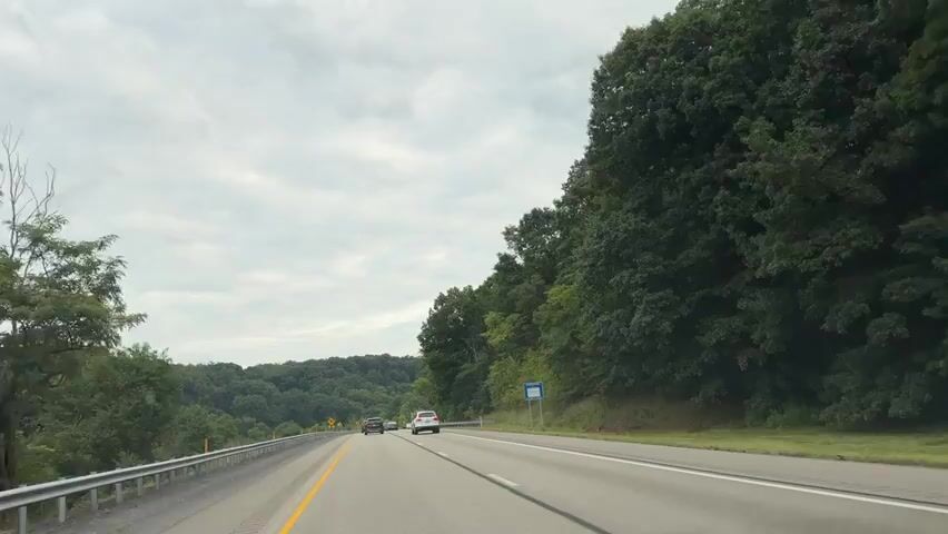 The video shows a POV shot from inside a vehicle driving on a highway. The highway has multiple lanes and a guardrail on the left side. The right side of the road is bordered by a steep, tree-covered hill. Several cars are visible driving on the highway ahead. In the distance, rolling hills covered in trees are visible. The sky is overcast. An orange dump truck passes the vehicle from the right lane.