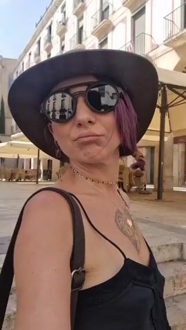 A medium shot shows a woman wearing a brown hat and sunglasses. She has purple hair and is wearing a black dress and a necklace. She is standing in front of a building with a dome and a colonnade. The woman is looking at the camera and smiling. The video is shot outdoors on a sunny day.